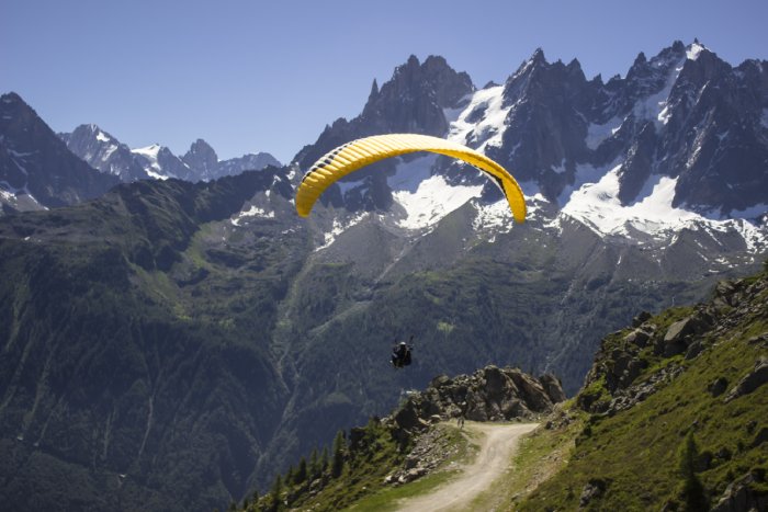 A paraglider sails high above an Alpine mountainside with snow-covered peaks in the background.