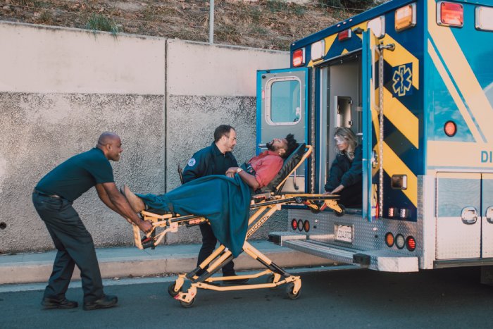 A man in a stretcher is wheeled out of an ambulance by three paramedics.