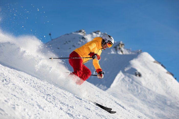 A smiling woman in yellow and red clothing skis on a steep slope in the high mountains on a sunny day.